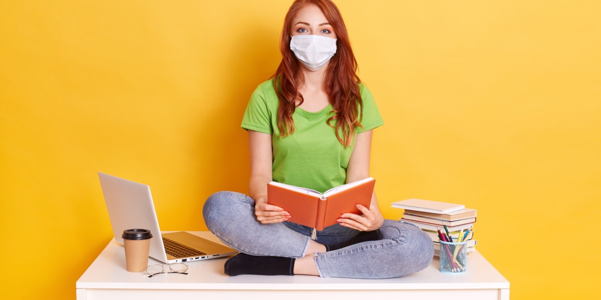 young-student-medical-mask-studying-home-while-quarantine-bored-distance-learning-sitting-with-crossed-legs-white-table-with-book-hands